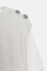Top de cuello redondo y manga corta con diseño de capas que combina una remera de algodón blanca con un chaleco de punto gris superpuesto. Presenta detalles de botones joya en el hombro.