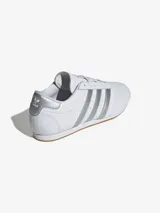 Zapatillas Adidas Taekwondo Lace blancas con detalles plateados en las tres tiras laterales y el talón. Diseño de perfil bajo con puntera pespunteada y suela de goma color caramelo.