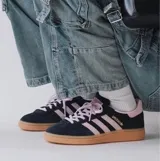 Championes Adidas Handball Spezial de corte bajo, confeccionados en gamuza negra con detalles en rosado en las icónicas tres tiras, cordones y talón. Presenta una suela de caucho color caramelo y el logo de la marca en dorado en el lateral.