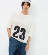 Remera de corte oversized confeccionada en tejido jacquard de color blanco con textura calada. Presenta mangas cortas, cuello redondo y un diseño gráfico deportivo con el número 23 en color negro en la parte frontal.