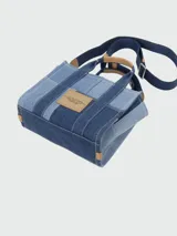 Cartera tipo tote bag de Marc Jacobs, confeccionada en denim con diseño de patchwork en diferentes tonos de azul. Presenta el logo estampado en el frente, doble asa superior y correa de hombro ajustable y extraíble en color azul marino con detalles en cuero tostado.