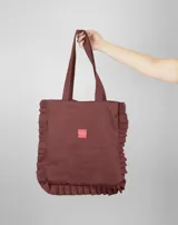 Tote bag color bordó con diseño de volados en los laterales y la base. Cuenta con doble asa larga para llevar al hombro y una etiqueta rectangular pequeña en el centro.