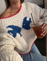 Sweater de tejido de punto grueso en color crema, con diseño de mariposas en color azul y detalles de terminación en color rojo en el cuello, puños y ruedo. Presenta un calce relajado, escote amplio y mangas largas.