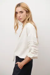 Camisa de color crudo con diseño romántico, cuello redondo con cordón ajustable terminado en borlas, y mangas tres cuartos abullonadas. Presenta detalles de bordados calados verticales en el frente y horizontales en las mangas.