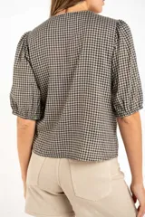 Blusa de algodón con estampado de cuadros vichy en tonos negro y blanco. Presenta escote en V, cierre frontal con botones y mangas cortas abullonadas con terminación elástica.