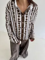 Camisa blanca de manga larga con delicados bordados florales verticales y detalles de encaje. Presenta cuello clásico, cierre frontal con botones y terminaciones festoneadas en los puños y el ruedo.