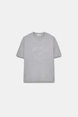 Camiseta gris de cuello redondo y manga corta, con un estampado en relieve que dice "NEW YORK CITY" y "BROOKLYN". El tejido tiene un sutil brillo.