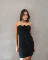 Vestido corto strapless negro con diseño drapeado en toda la prenda, detalle de aplique floral en el centro del escote y caída en cascada con volados frontales. Presenta pequeños puntos brillantes distribuidos por toda la superficie.