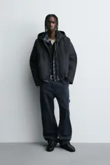 Cazadora tipo bomber de denim color negro con efecto lavado, corte boxy fit, cierre frontal de cremallera, capucha ajustable con cordón y bolsillos laterales de vivo.