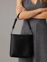 Cartera tipo saco pequeña Calvin Klein color negro, de cuero sintético granulado. Tiene dos correas desmontables: una correa fina ajustable y una correa ancha ajustable con logo jacquard.
