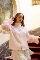 Camisa de lino color rosa pálido, con cuello alto decorado con volados, cierre frontal con botones forrados y mangas largas amplias con puños terminados en volados.