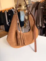 Cartera tipo hobo de textura gamuzada en color marrón oscuro, con diseño flexible y asa larga regulable. Presenta un detalle de flecos largos desmontables en uno de los laterales y cierre superior.