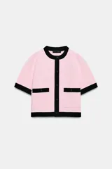 Chaqueta de punto rosa con ribetes negros en el cuello, la botonera, los bolsillos, los puños y el ruedo. Cuenta con manga corta, cuello redondo, bolsillos frontales de plastrón con botones y cierre frontal abotonado.