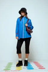 Camisa azul de corte oversize, con cierre frontal de botones y detalles de cintas onduladas en colores contrastantes a lo largo de la botonadura y en los puños. Presenta cuello clásico y mangas largas.