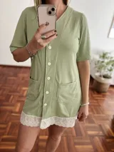 Camisola o vestido corto de tejido acanalado color verde claro, con cuello en V, cierre frontal de botones blancos y dos bolsillos frontales tipo parche. El ruedo está adornado con un borde de encaje blanco.