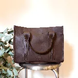 Cartera de cuero color tostado, estructurada, con doble asa de mano, correa desmontable y dos bolsillos frontales verticales con cierre. Presenta detalles de costuras pespunteadas y el logo de la marca grabado en la parte inferior.