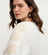 Vestido corto de playa confeccionado en voil de algodón BCI color blanco, con escote en V con lazo, cutouts laterales, mangas largas y bordados de caracoles de mar.