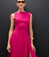 Vestido midi de sastrería color fucsia, sin mangas y con escote redondo. Presenta un adorno metálico dorado en uno de los hombros y una abertura lateral en la falda que aporta movimiento.