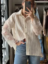 Camisa blanca de manga larga con diseño texturizado en el cuerpo y mangas de encaje con motivo floral translúcido, cuello camisero y cierre frontal con botones.