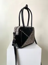 Cartera rígida negra con detalles en rojo y tachas plateadas. Tiene herrajes y cierre metálico plateado marca YKK, asa fina para llevar al hombro o en la mano y correa larga extra regulable. El interior es forrado, tiene un bolsillo con cierre enmarcado en cuero, y un segundo bolsillo plaqué.
