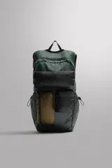 Mochila deportiva de tejido técnico en color verde oscuro, con múltiples bolsillos frontales en tonos negro y marrón. Cuenta con compartimento principal con cierre termosellado, bolsillos laterales ajustables con cordón, parte posterior acolchada y asas ajustables con hebilla de liberación rápida.