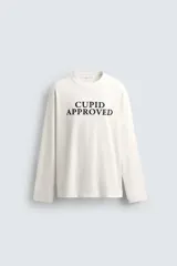 Camiseta de manga larga color blanco roto, corte relajado (relaxed fit) con cuello redondo y texto estampado en negro en el pecho que dice "CUPID APPROVED".