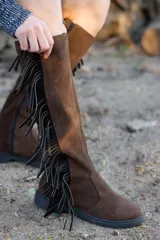 Botas de caña alta confeccionadas en cuero nobuk color chocolate, con detalle de flecos laterales en color negro, suela plana y cierre lateral.