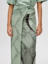 Pareo largo de viscosa en color verde menta, con diseño estampado que presenta gráficos abstractos y una figura central destacada en negro. Es una prenda liviana y fresca, ideal para usar sobre el traje de baño o como falda envolvente.