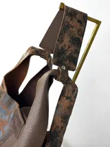 Cartera tipo hobo de cuero genuino con diseño bicolor que combina paneles de cuero liso marrón con paneles de cuero texturizado con efecto desgastado en tonos negro y marrón. Incluye un sobre extraíble a juego con cierre y etiqueta de la marca.