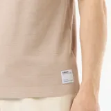 Camiseta de cuello redondo y manga corta color beige, con textura de nido de abeja y pequeña etiqueta decorativa en el borde inferior.