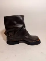Bota de caña media confeccionada en cuero con efecto desgastado, presenta un diseño holgado en la parte superior y una tira con hebilla metálica ajustable en el tobillo. Cuenta con suela de plataforma y taco cuadrado robusto.