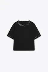 Top negro de manga corta con cuello redondo, adornado con perlas blancas aplicadas en el escote y en los puños de las mangas. Cierre posterior con botones de perla.