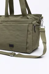 Cartera tipo tote bag de nylon color verde militar, con diseño amplio y liviano. Cuenta con asas superiores reforzadas, correa larga regulable y desmontable, bolsillos frontales y laterales con cierre metálico, y un pequeño aplique con logo en la parte inferior.