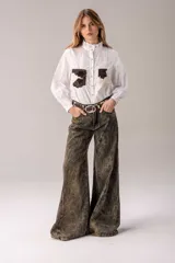 Camisa blanca de corte clásico con cuello alto tipo victoriano con detalle de puntilla. Presenta bolsillos frontales aplicados con textura de cuero con pelo en estampado animal print vacuno. Posee cierre frontal con botones y mangas largas con puños estructurados.