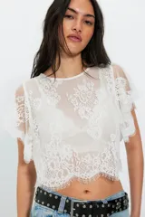 Top corto de encaje blanco con escote redondo, mangas cortas amplias con volados y ruedo festoneado.