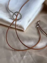 Choker marrón de gamuza con dije de doble aro entrelazado, uno liso y otro con incrustaciones de strass. Los extremos del choker tienen detalles de cuentas doradas.