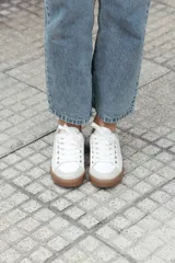 Zapatilla de estilo urbano en tonos marfil, con detalles en color dorado en el talón y ojales metálicos. Presenta suela de goma color caramelo y cordones al tono.