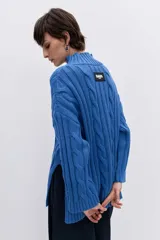 Sweater de punto con diseño de trenzas, cuello alto y mangas largas con puños acampanados. Presenta un corte holgado con terminación asimétrica en el ruedo.
