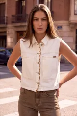 Chaleco de denim blanco sin mangas, con cuello camisero, bolsillos frontales plaqué con solapa y cierre frontal de botones con detalles de costuras onduladas en contraste.