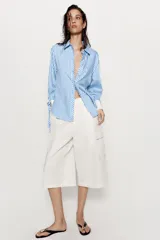Camisa de cuello solapa y manga larga con rayas verticales celestes y blancas. Cuenta con puños anchos blancos contrastantes, ajustables con hebilla dorada y lazos, y cierre frontal con botones dorados.