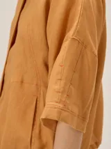 Chaqueta de lino y rayón color tostado, de corte holgado, mangas cortas amplias, cuello mao y cierre frontal oculto con cremallera. Presenta pespuntes a contratono en las costuras y cordón ajustable en el bajo.