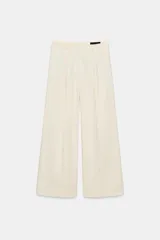 Pantalón wide leg de tiro medio color beige, confeccionado en hilatura de algodón. Presenta cintura con trabillas, bolsillos laterales ocultos en costura, detalle de pliegues en el delantero y cierre frontal con cremallera, botón y ganchos metálicos.