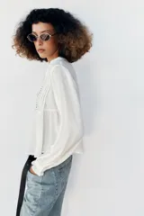 Blusa blanca de manga larga con cuello en V, cierre frontal con botones y detalles de bordados florales y encaje en el frente.