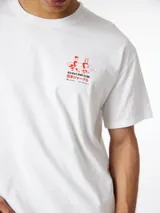 Camiseta blanca de algodón orgánico, con corte oversize, cuello redondo y manguita corta. Presenta un pequeño estampado rojo en el lateral superior izquierdo con la frase 'Go Out And Slide' y caracteres en japonés junto a una ilustración de dos personas cargando una tabla de surf.