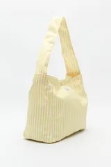 Bolso tipo tote bag de algodón, color amarillo pálido con rayas verticales blancas. Tiene un asa ancha para llevar al hombro y un pequeño parche con texto en el frente.