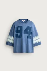 Camiseta boxy fit de color azul con efecto lavado vintage, cuello redondo y mangas por debajo del codo. Presenta un gran número '94' estampado en el frente y franjas horizontales en las mangas.