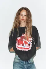 Camiseta de corte oversize con efecto lavado, cuello redondo y manga corta. Presenta una estampa gráfica central que representa una figura femenina sobre un pastel decorado con cerezas.