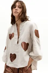 Camisa oversize de lino color beige, con cuello mao, cierre frontal con botones de nácar y mangas abullonadas con puño elástico. Presenta apliques de corazones en material metalizado tono bronce.