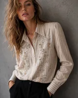 Camisa de corte clásico a rayas verticales en tonos neutros, con cuello camisero y cierre frontal de botones. Presenta un delicado bordado floral con canutillos y lentejuelas brillantes en el frente.