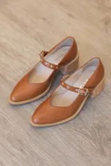 Zapato Mary Jane de cuero marrón con escote en V, punta redondeada, taco ancho efecto madera y tira en el empeine con hebilla metálica y detalles de ojalillos.